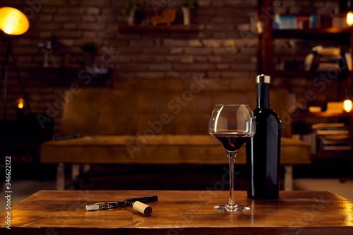 Wine glass and a bottle of red wine on table at home, cosy dark living room in background. Autumn mood with dark and warm colors.