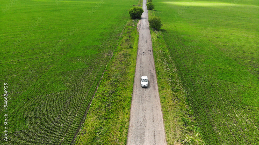 Local tourism concept. White car driving along the old asphalt road between two spring agricultural fields. Aerial. Local tourism concept. White car driving along the old asphalt road between two spring agricultural fields. Aerial.