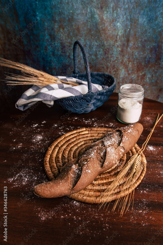 Handmade rye bread over a wooden table with spikes and cloth in a basket and a flour jar.