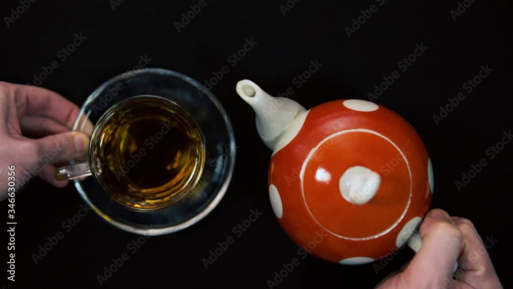 Tea ceremony. A male hand cleans up hot glass red polka dot ceramic teapot with flowers next to glass cups for a bowl. Black background, oncoming light 4k Tea ceremony. A male hand cleans up hot glass red polka dot ceramic teapot with flowers next to glass cups for a bowl. Black background, oncoming light 4k