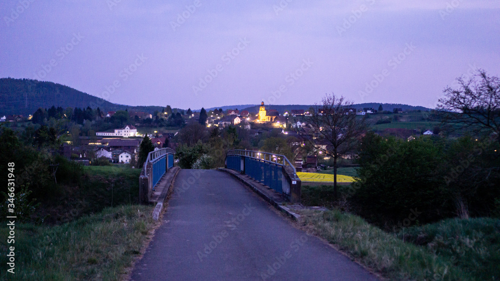 Fototapeta Panorama von einem kleinen Dorf am Horizont mit herausstechendem Kirchturm bei Nacht. Lila, blauer Himmel und dunkle Farbtöne. Kirchturm leuchtet gelb. Brücke bzw. Weg im Vordergrund. Frankenwald.