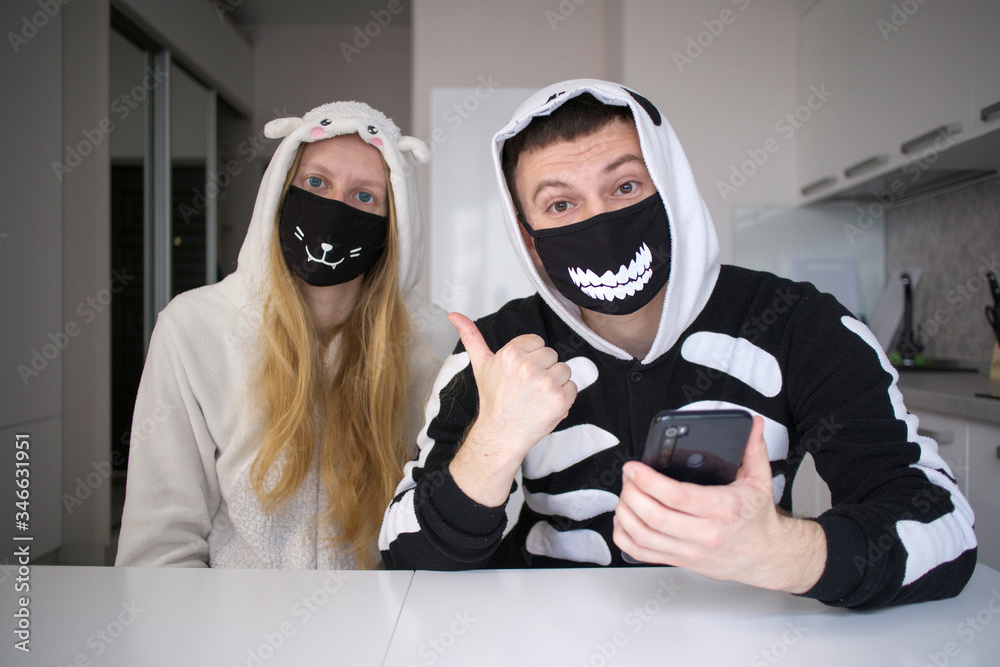 young brunette guy in black with white pajamas with a hood (kigurumi) and a blonde girl in white pajamas (kigurumi) in unusual black masks with a white print have fun on self-isolation in the kitchen