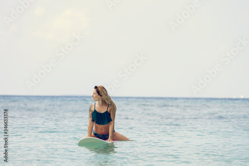 Beautiful surfing girl in bikini and with wet hairs sitting on a front of big longboard surf board and waiting for the wave in open ocean water. Modern active sport lifestyle and summer vacation.