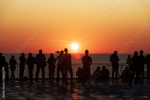 Silhouettes of people against the setting sun on deck of a cruise ship. Calm ocean and clear orange sky in evening time. Cruise vacations background