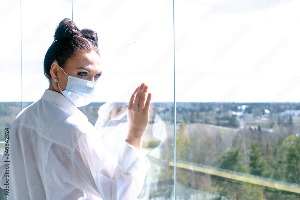 Fototapeta premium In a medical mask looks out the window. Concept: self-isolation, virus, coronavirus. be at home
