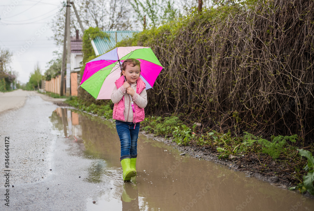A beautiful little girl walks through puddles with a colorful umbrella and green rubber boots. Horizontal orientation. Walk a cute girl in the fresh air after the rain.
