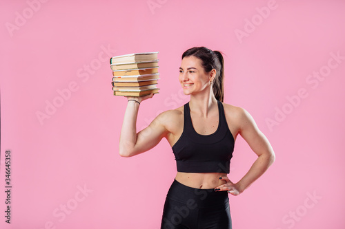 Fototapeta Naklejka Na Ścianę i Meble -  Beautiful, attractive, muscular girl holding heavy set of books on one hand, and with her second hand showing her large muscles. Demonstrating pumped up body in photo studio on pink background
