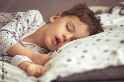 Little blond boy sleeping in his bed. Restless sleep of a baby with a cold. The kid sleeps with his mouth open. My little boy sleeping by the light of the window. photo