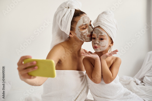 A young caucasian mother and little daughter with wrapped hair in white bath towels with a mask on their faces have a fun and make selfies on the phone