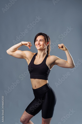 Portrait of a happy elegant caucasian brunette woman showing her biceps muscles on the arms in black clothes and fitness over gray background, smiling and looking at camera