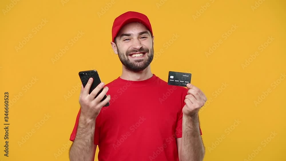 Delivery guy man in red cap t-shirt workwear hold in hand credit bank card using cell phone isolated on yellow background studio Professional male deliveryman employee working courier Service concept Delivery guy man in red cap t-shirt workwear hold in hand credit bank card using cell phone isolated on yellow background studio Professional male deliveryman employee working courier Service concept