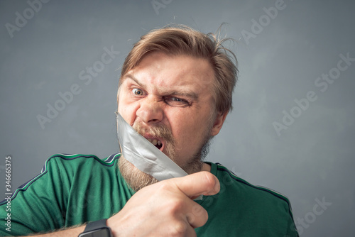 Close-up portrait of a bearded man ripping the tape off his face. The concept of what can't be said is censorship.