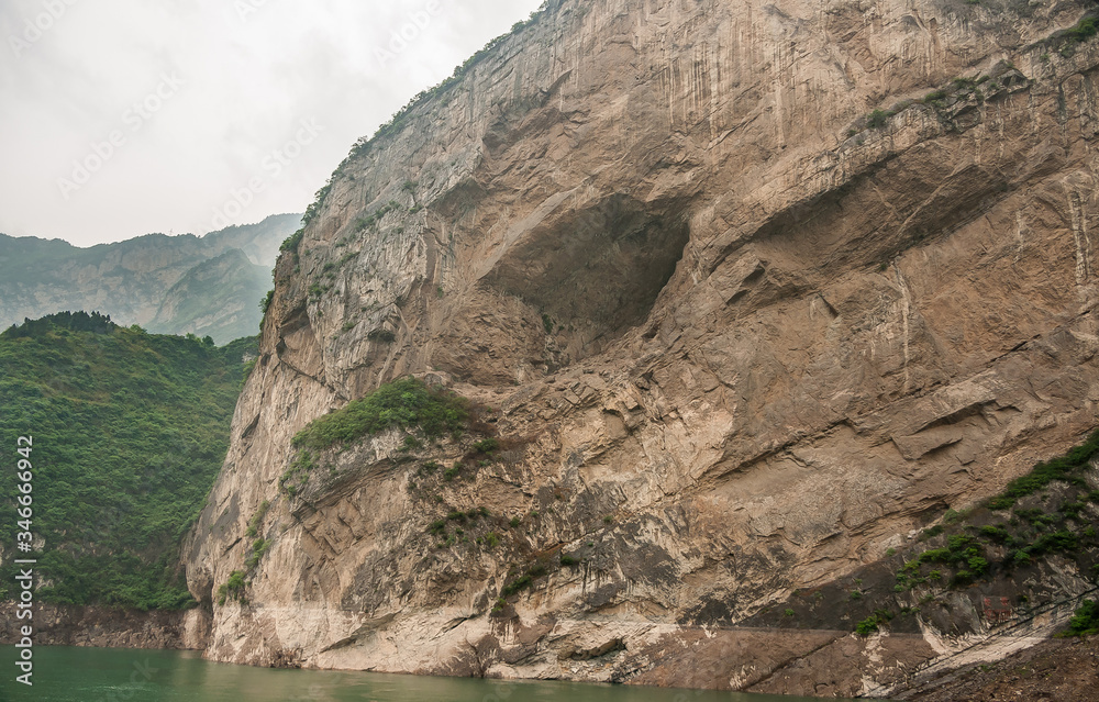 Xiangxicun, China - May 6, 2010: Xiling gorge on Yangtze River. Large cave hole in brown rock cliff descending in green water. Green covered mountains as backdrop. Patch of silver sky.