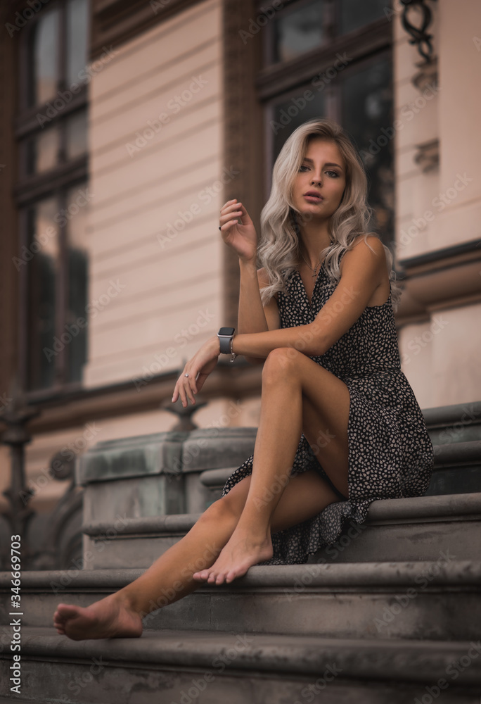 Naklejka premium Beautiful blonde young girl wearing fashionable clothes sitting on the stairs in a blue sverfan bassek against the background of a light building