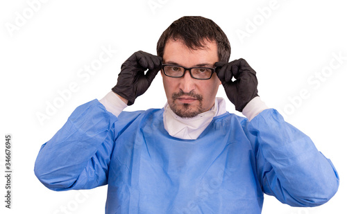 Portrait of male doctor in protection clothing and disposable black gloves, putting the eyeglasses, isolated on white background. His offers healthcare services and treats patients with Coronavirus © Kindarts