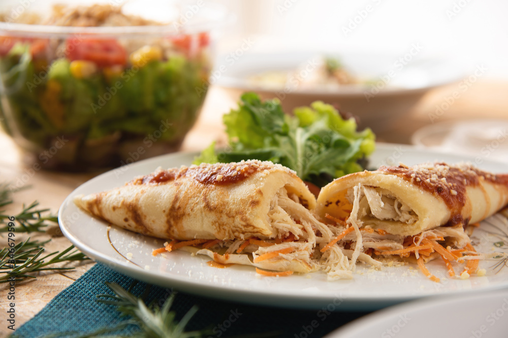 Chicken pancake on top of a white plate on a wooden table with salads in the background. Brazilian pancake