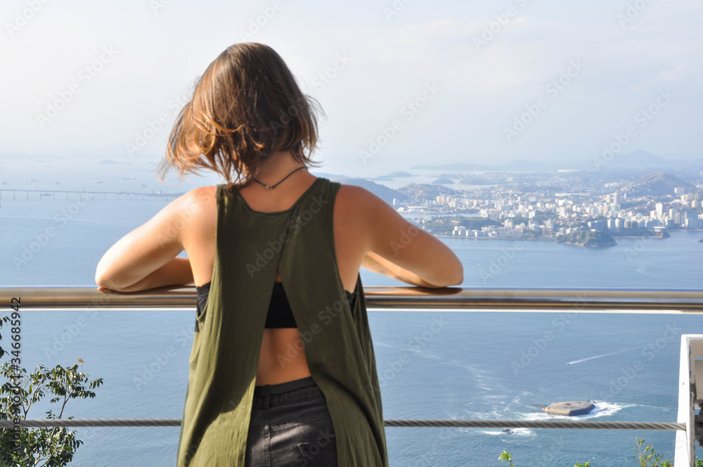 A young woman is standing at a sightseeing point looking at the landscape from the sea and the city. She is wearing a green shirt.