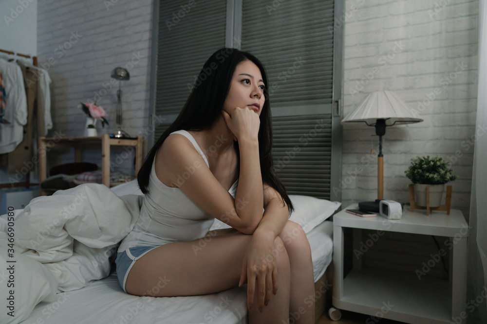 Young asian chinese woman sitting on white comfort bed in dark bedroom while wake up in early morning. girl suffering from depression in late night. pensive lady looks thoughtful looking out window. Young asian chinese woman sitting on white comfort bed in dark bedroom while wake up in early morning. girl suffering from depression in late night. pensive lady looks thoughtful looking out window.