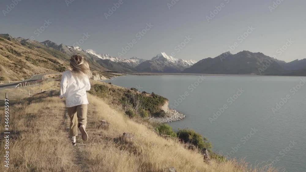 Blonde girl running beside a blue lake with snowy mountains in the background during sunrise in slow motion. Blonde girl running beside a blue lake with snowy mountains in the background during sunrise in slow motion.