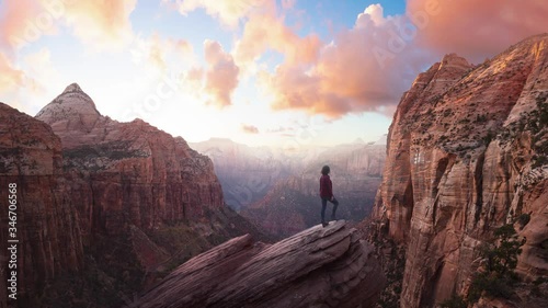 Adventurous Woman at the edge of a cliff is looking at a beautiful landscape view in the Canyon during a vibrant sunset. Taken in Zion National Park, Utah, United States. Parallax Panorama