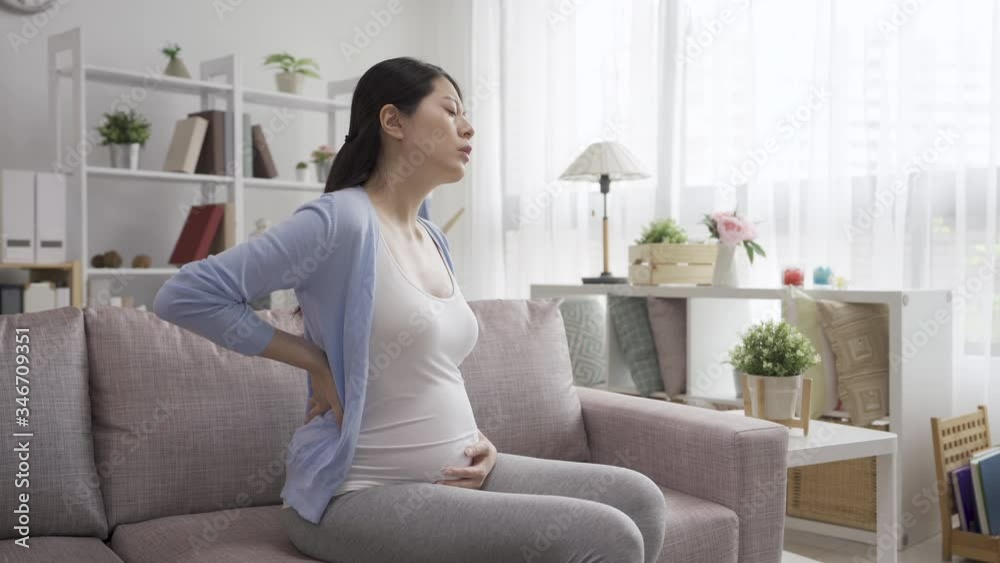 Young pregnant woman suffering from backache at home living room. frowning future motherhood lady sitting on sofa with painful back and massage it. female hands touching big belly with expecting baby Young pregnant woman suffering from backache at home living room. frowning future motherhood lady sitting on sofa with painful back and massage it. female hands touching big belly with expecting baby