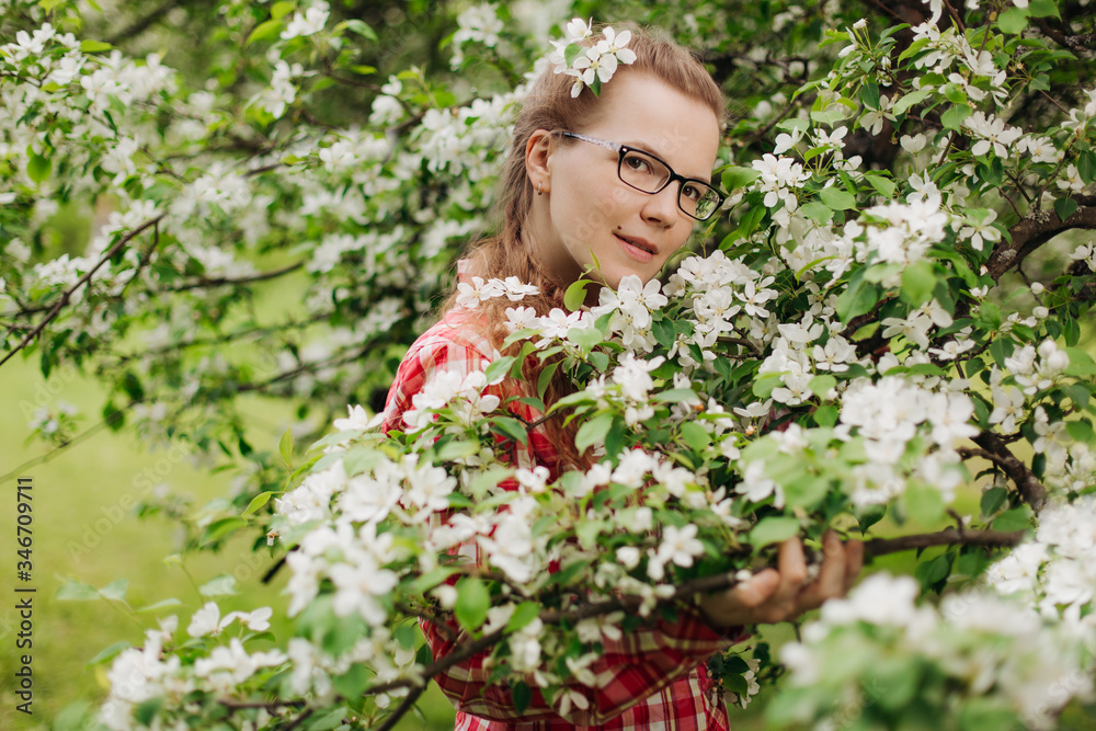 Beautiful young happy woman with glasses in a flowered garden. Girl among the flowers of apple trees, cherries. Springtime scene. Positive model in a casual style. Lifestyle Beautiful young happy woman with glasses in a flowered garden. Girl among the flowers of apple trees, cherries. Springtime scene. Positive model in a casual style. Lifestyle