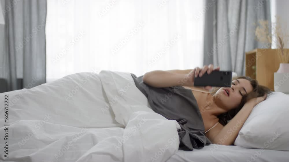 Young brunette woman waking up at home, looking at smartphone screen and getting worried. She sitting on bed and start typing message in the morning Young brunette woman waking up at home, looking at smartphone screen and getting worried. She sitting on bed and start typing message in the morning