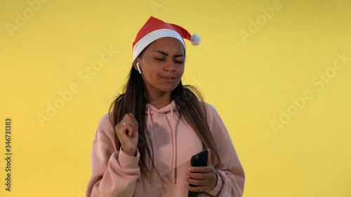 Happy african woman in Santa hat and wireless headphones listening to the music on yellow background. She singing some Christmas songs.