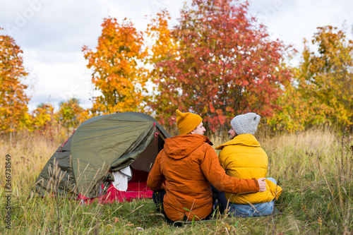 travel, love, autumn and trekking concept - back view of couple in love sitting near green tent in autumn forest