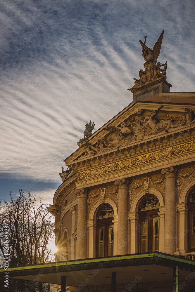Obraz premium Beautiful opera house in Ljubljana, slovenia during sunset. Detail of the front facade with sun just sending rays of light at the side wall. Nice cloud texture above the house.