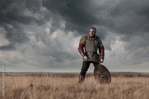 Warrior Viking in full arms with axe helmet and shield on dark sky background. Medieval scandinavian berserk red beard and fur costume attacks enemy. Concept historical photo