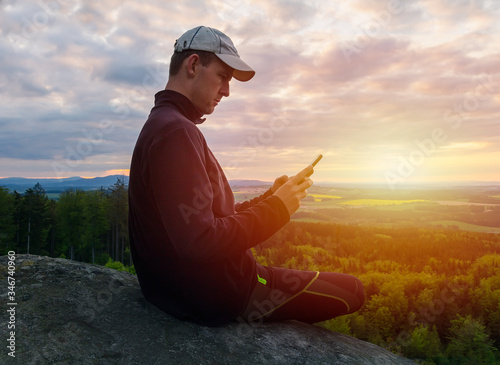 Young caucasian man sitting on rock and using his mobile at sunset. Czech republic