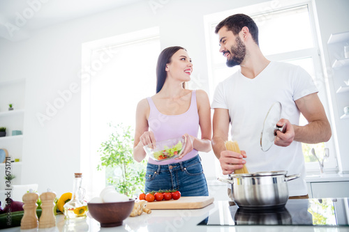 Low angle view photo of two people married spouses chef prepare veggie dish lunch woman mix fresh salad man boil macaroni stove in kitchen house indoors