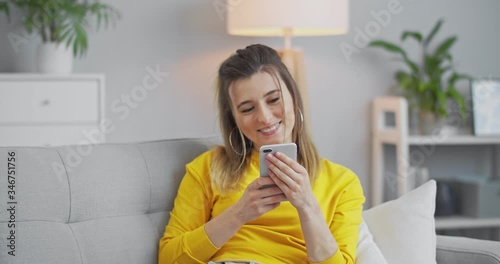 Relaxed young woman holding smart phone looking at cellphone screen laughing enjoying using mobile apps for shopping having fun playing games chatting in social media sitting on couch at home. photo