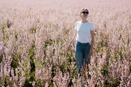 Young woman with brown hair in white T-shirt and blue jeans poses in a pink sage field. Warm midday light.