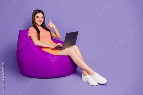 Full body profile photo of nice lady sitting comfy beanbag browsing notebook raise thumb finger up wear orange striped t-shirt jeans mini skirt isolated pastel purple color background