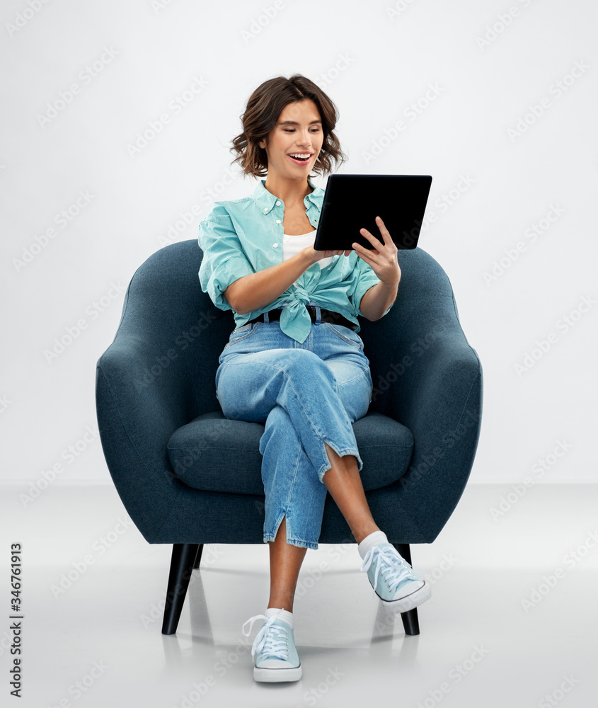 people and technology concept - portrait of smiling young woman in turquoise shirt and jeans with tablet pc computer sitting in modern armchair over grey background