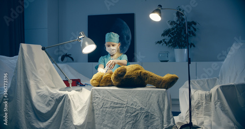 Cute little girl wearing doctor suit playing at home, pretending to be a surgeon, providing medical care to her teddy bear