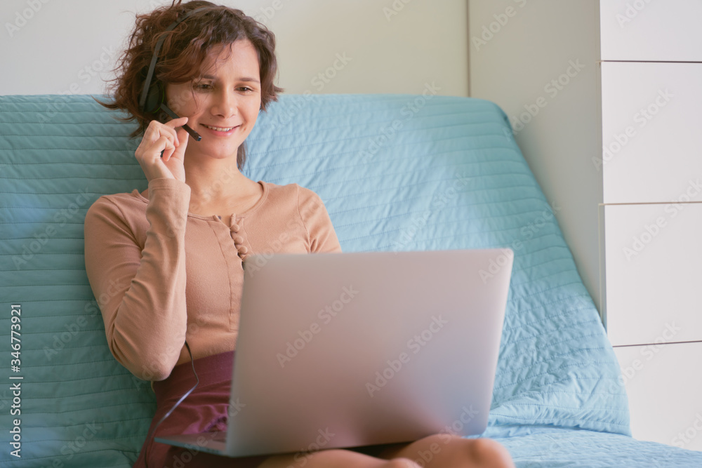 Fototapeta premium Young specialist works from home, communicates via headset, consults online, solves important questions problems of clients. Lady beautiful smile wavy hair Bob is wearing shirt skirt laptop is on lap