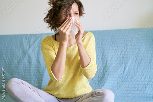 Young woman during coronavirus pandemic, sneezes into napkin, runny nose. Sick female suffers from flu, is treated in an apartment, freelancer with wavy Bob hairstyle is covered with blanket sofa 