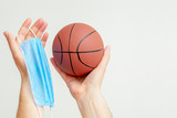 Basketball ball with medical protective mask in man's hands on light background during coronavirus with copy space. Basketball on pause due to coronavirus.