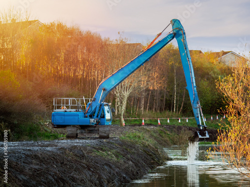 Blue excavator working on a river  Cleaning bottom. Warm sunny day  Cloudy sky  Heavy equipment in use concept.