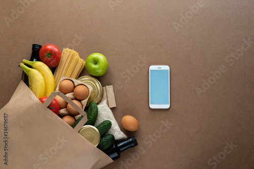 Paper bag with food supplies and mobile phone on neutral beige background. Food delivery, mobile app for quarantine, donation, coronavirus concept. Copy space for text, mockup