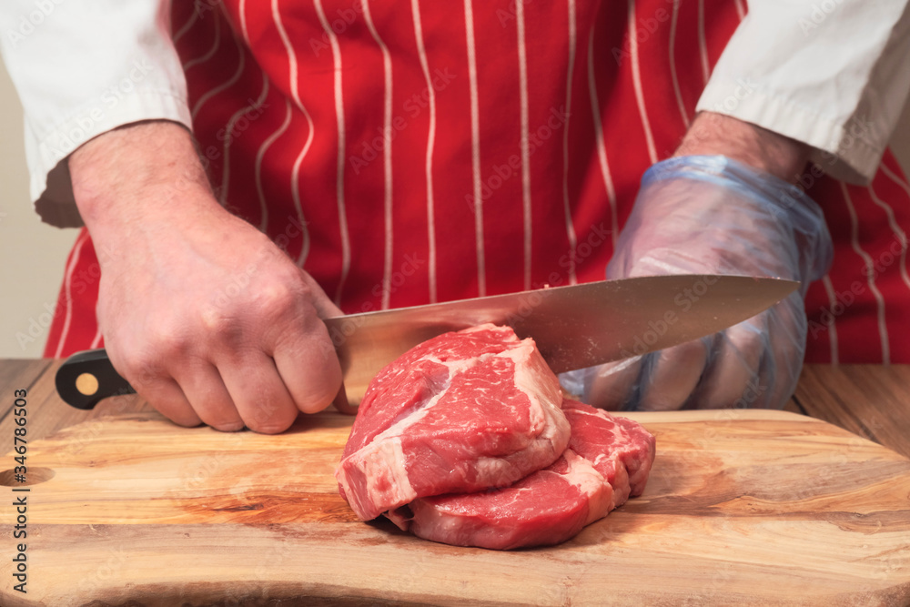 Two fresh rib eye steaks on a wooden cutting board. Professional butcher in red apron holding knife in one hand, other hand in blue plastic glove. Meat industry concept
