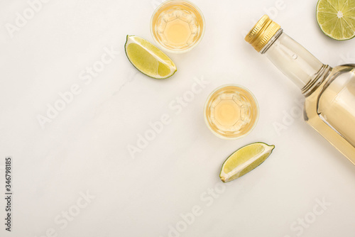 top view of golden tequila in bottle and shot glasses with lime on white marble surface