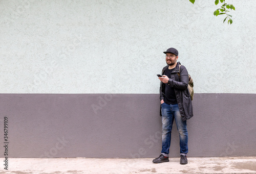 a man in a black cap, light summer raincoat and glasses holds a phone in his hands. a man in jeans. the man is happy and smiles. two-color background © Илья Антропов
