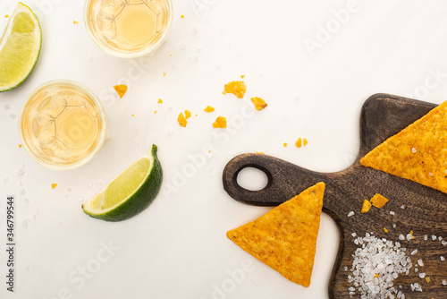 top view of golden tequila in shot glasses with lime, salt and nachos on wooden cutting board on white marble surface