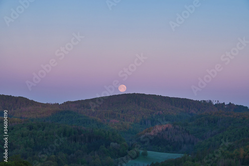 Morgendämmerung bei Vollmond mit Panorama Landschaftsansicht mit Wald und bunten Farben am Himmel photo