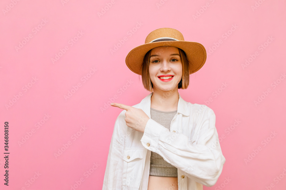 Happy attractive girl stands against a pink background in a hat and white summer clothes,looks in camera and points finger to the side. Smiling beautiful lady in white shirt points to copy space Happy attractive girl stands against a pink background in a hat and white summer clothes,looks in camera and points finger to the side. Smiling beautiful lady in white shirt points to copy space
