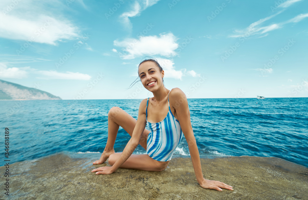 smiling woman in swimsuit sitting on stone beach, enjoying sea ocean view and blue sky. concept summer sea vacation and travel
